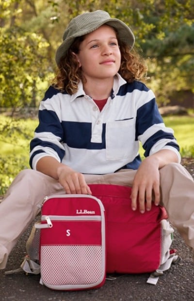 A child in a striped shirt and hat sits outdoors with a red L. L. Bean backpack and red lunch box.