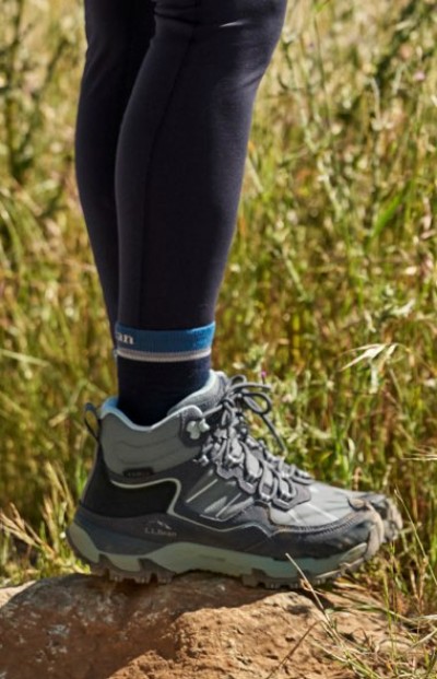 Gray hiking boots worn with black leggings on a dirt trail with tall grass in the background.