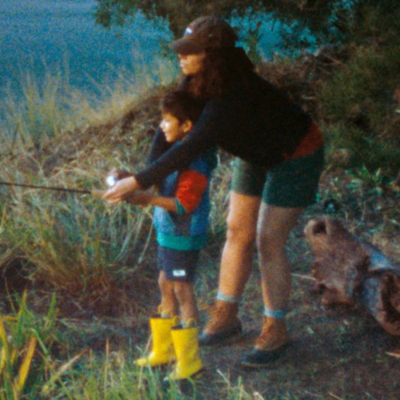 A mom behind her young son, arms over his shoulders, helping him with his fishing rod.