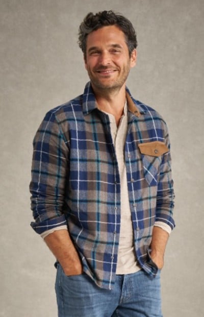 Man in blue and gray plaid shirt with rolled-up sleeves stands against gray background with hands in pockets.
