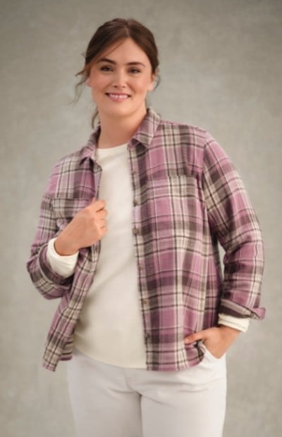 Woman in purple plaid shirt over white top stands against plain background with one hand in pocket.