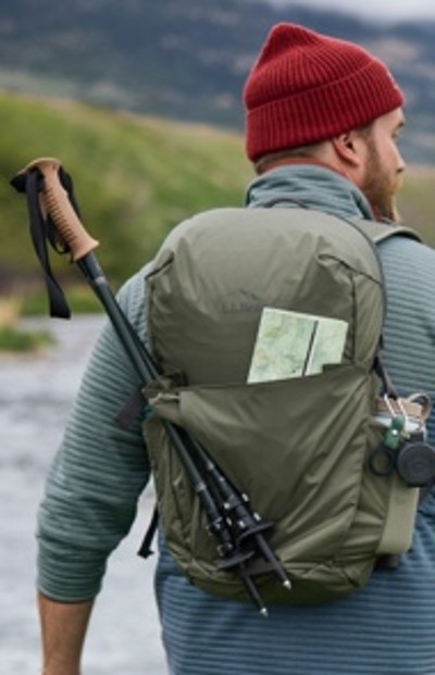 Person in red beanie and blue jacket carries green backpack with trekking poles, map, and gear attached.