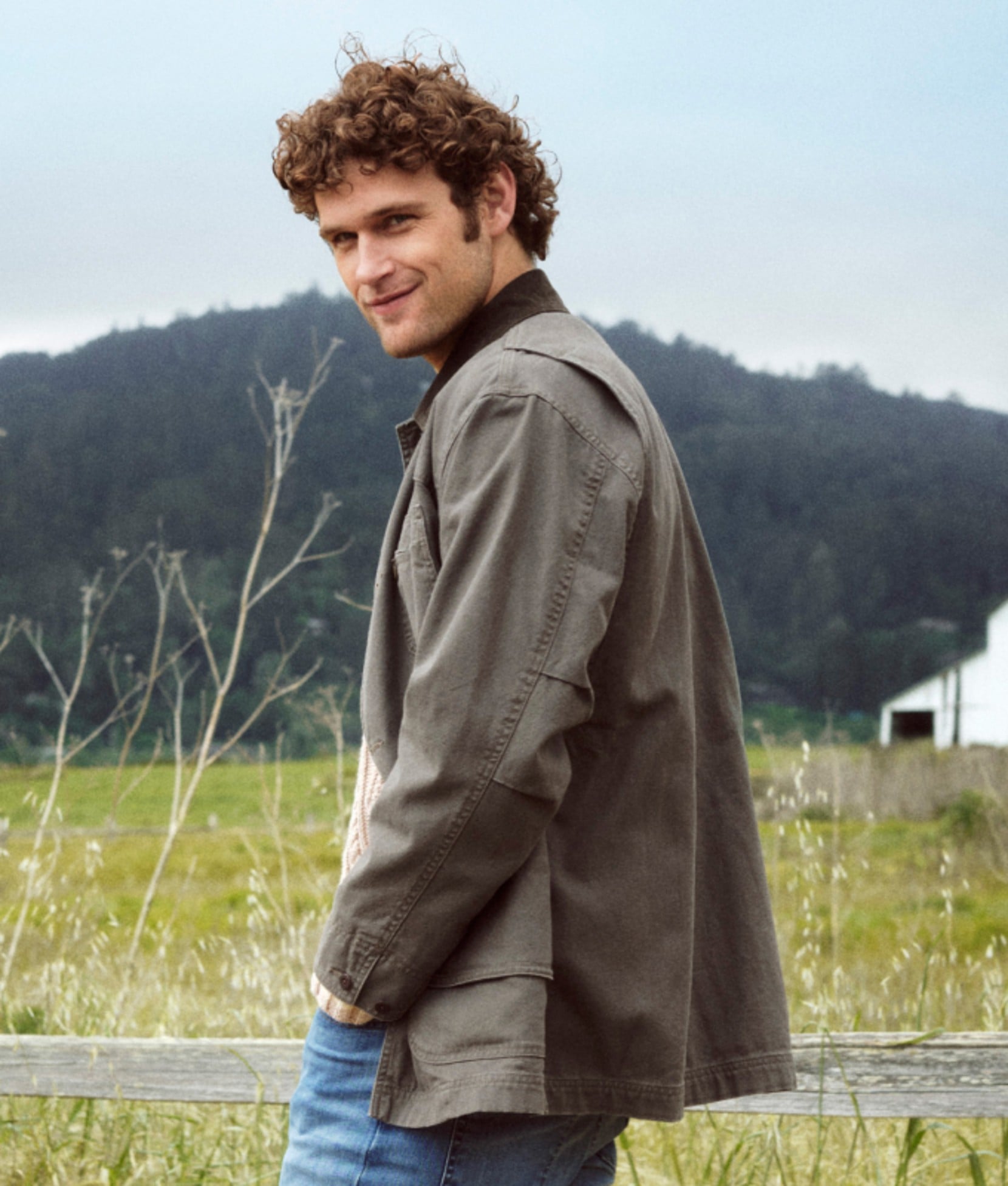 Man wearing an olive field jacket standing in a grassy outdoor setting. 