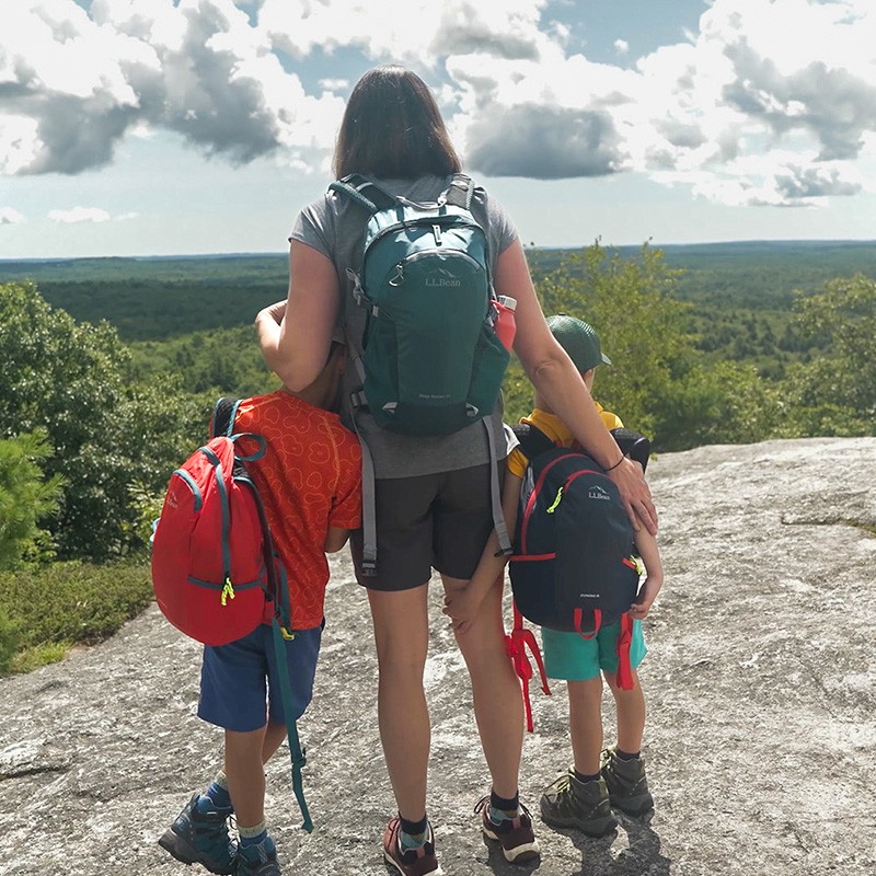 A mom and two children with backpacks stand on a rocky surface, overlooking a green landscape under a partly cloudy sky.