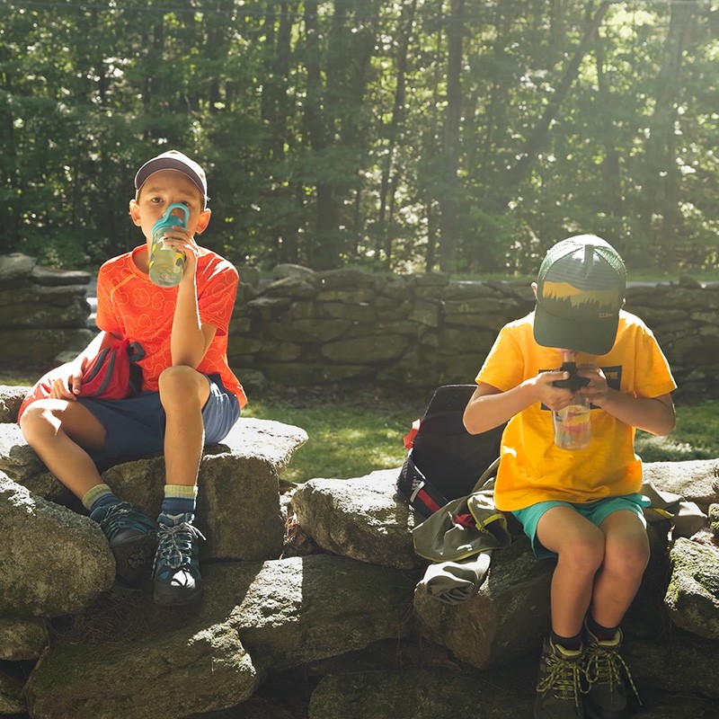 Two boys sitting on a stone wall in a forest drinking from water bottles.