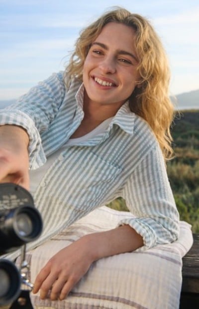 Reclining outdoors on a cushion, the person reaches toward a camera while wearing a striped long‑sleeve shirt.