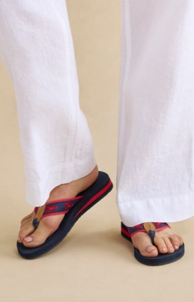 Navy flipā€‘flops with red straps worn with loose white pants, shown against a simple beige background.