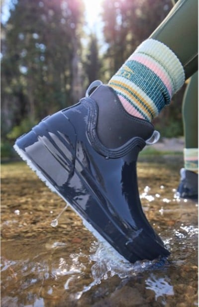 Dark blue waterproof shoe splashing through a shallow stream, paired with striped socks in a forest setting.