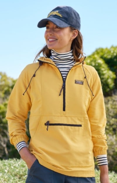 A woman in yellow jacket, striped shirt, and blue cap stands outdoors with green foliage and trees in the background.