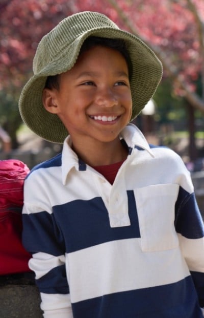A boy wearing a green hat and a white and blue striped shirt leans against a stone wall on a sunny day.