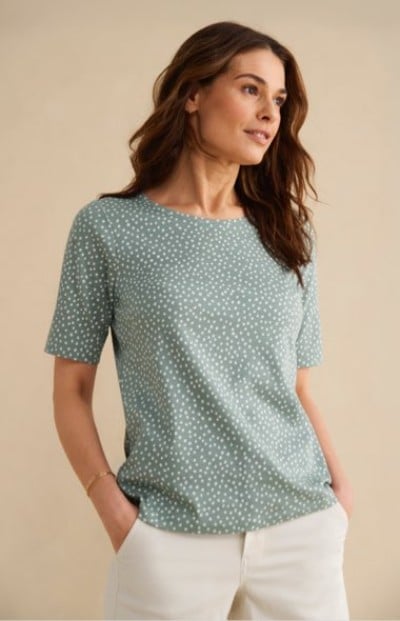 Wearing a patterned short‑sleeve top and light pants, a person stands with hands in pockets against a neutral background.