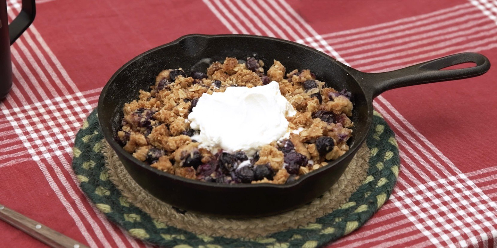 A skillet with blueberry cobbler topped with a dollop of whipped cream on a tablecloth.