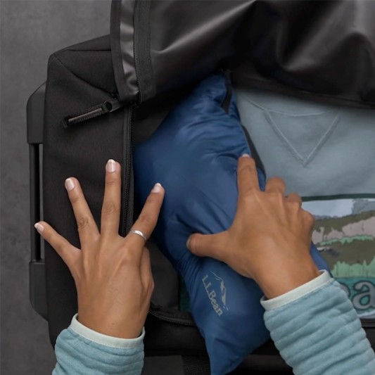 Hands place a blue stowawayed jacket into a black duffle bag.