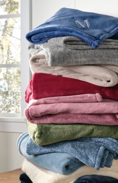 Stack of colorful fleece blankets in shades of blue, gray, red, pink, and green sits by a window, with one monogrammed “M.”