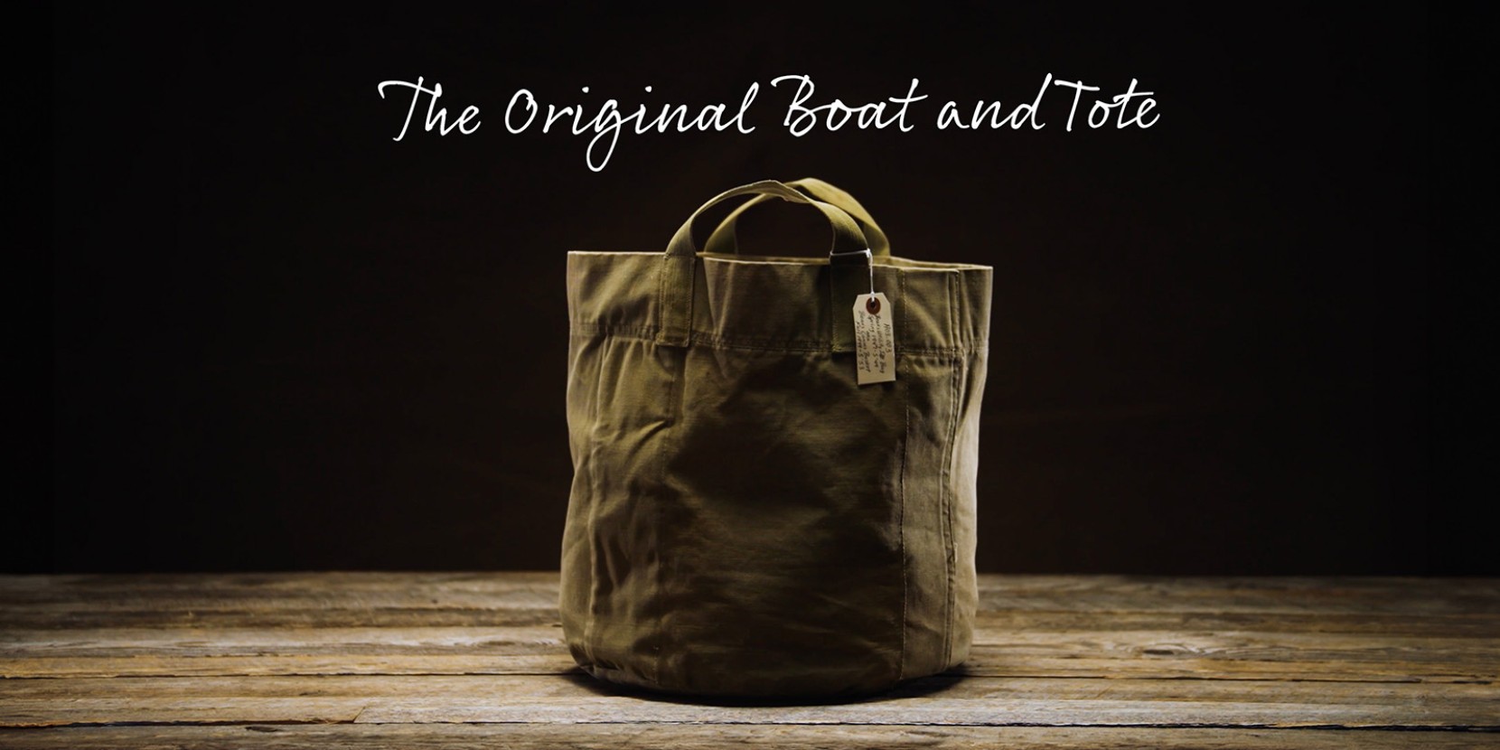 A khaki tote bag with handles on a wooden surface; text above reads “The Original Boat and Tote.”