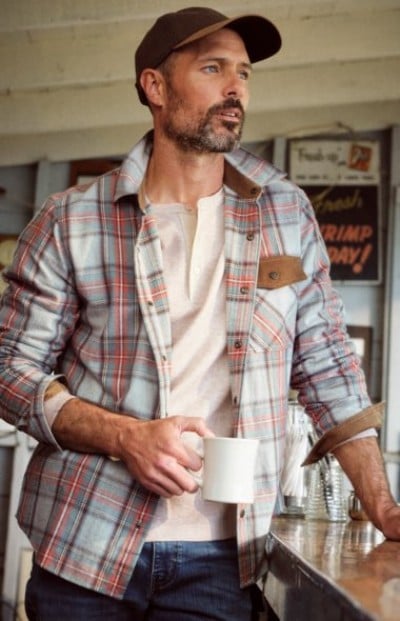 Person holding a mug on a porch, dressed in a plaid overshirt, baseball hat and jeans.