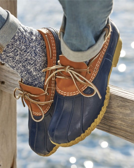 Close up image of someone wearing Bean Boots sitting a fence with the ocean in the background.