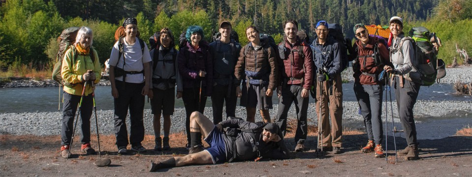 A group of 11 people standing by a river wearing backpacks and hiking gear.