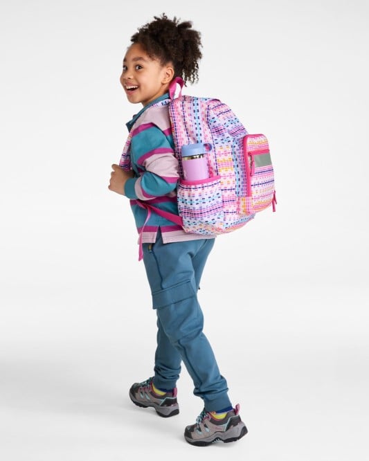 Smiling child in casual clothes walking and looking back, wearing a colorful backpack with side pocket items visible.