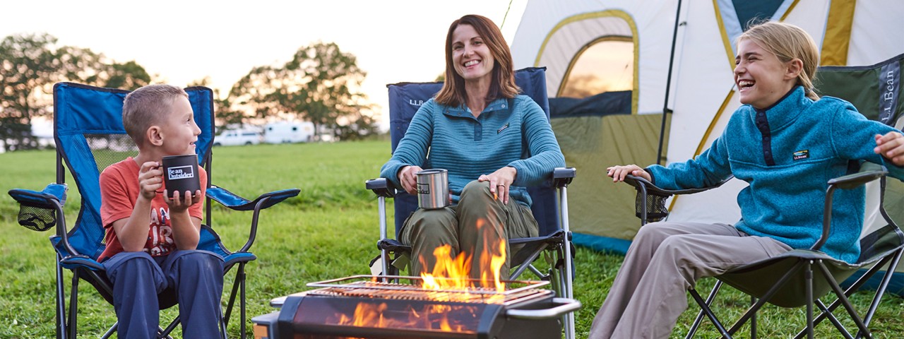 A woman and two children sit by a fire in front of a tent.