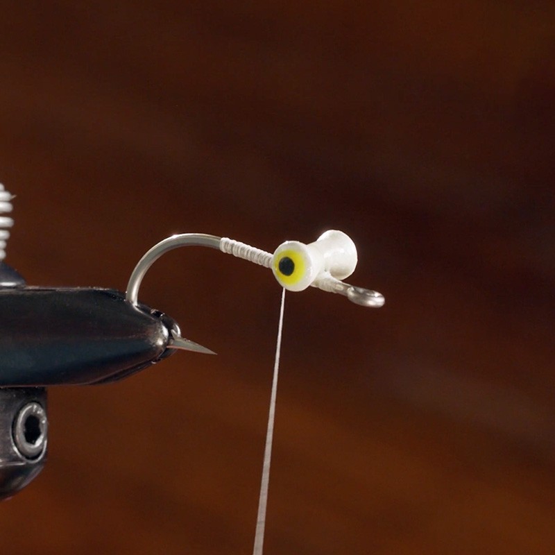 A close-up of a fishing hook being tied with white thread, featuring a yellow and white bobber attached to the hook.