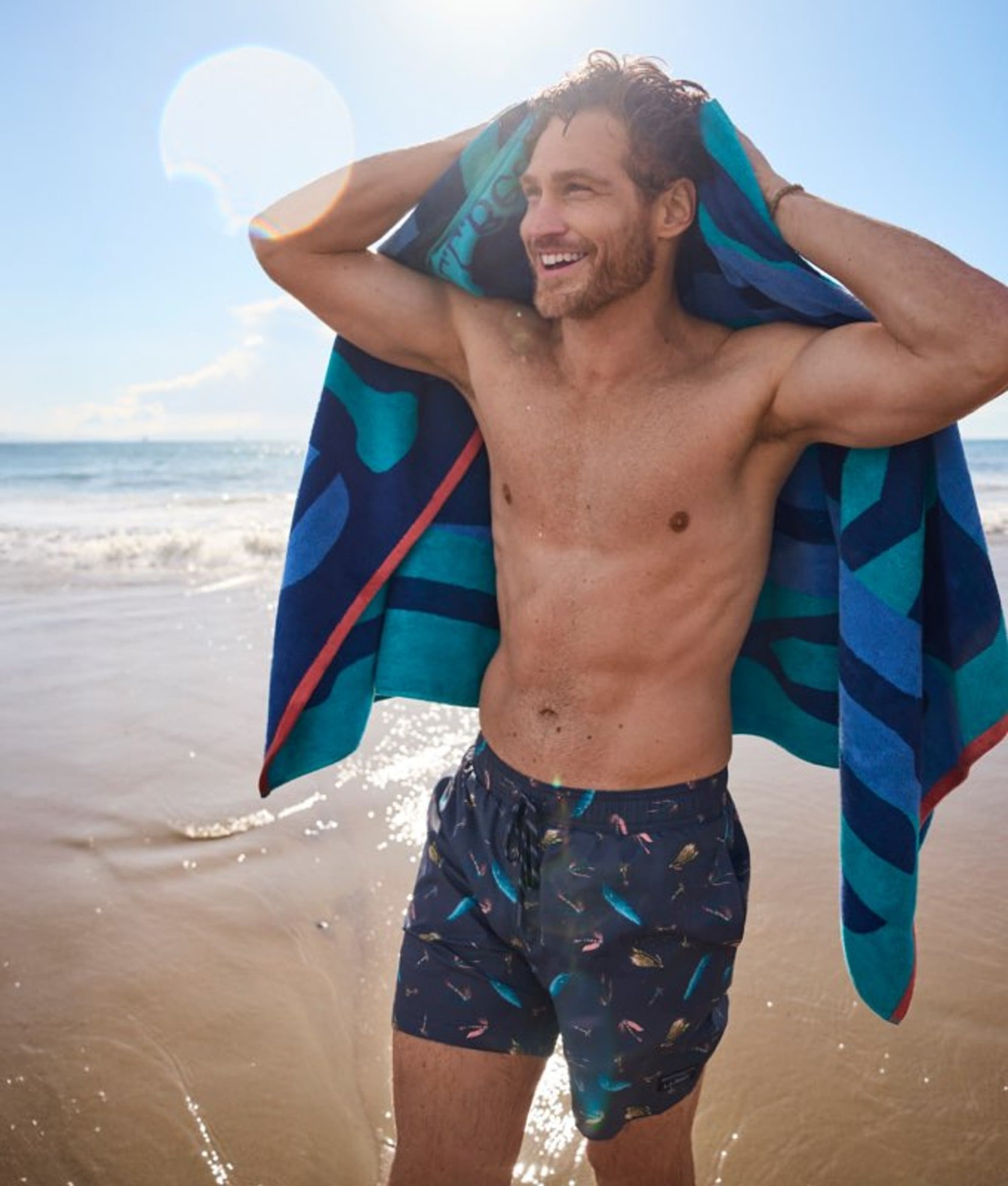 Man in blue swim trunks drying off with blue and teal beach towel on beach.