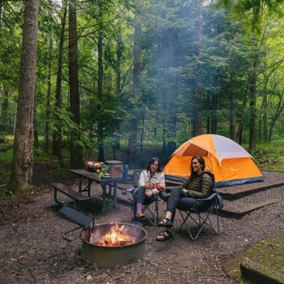 2 women sitting in camp chairs at their campsite, fire roaring, tent in the background.