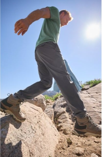 Person stepping across sunlit rocks in outdoor clothing and rugged hiking shoes under a bright blue sky.