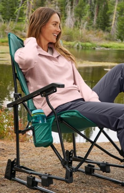 Person relaxing in a green outdoor chair beside a calm lakeside, wearing a light pink top and dark pants.