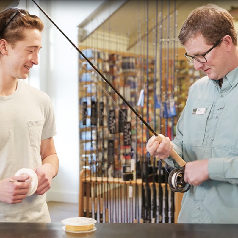 Two people in a fishing store, one holding a fishing rod and the other holding fishing line.