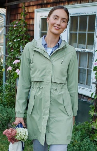 Person wearing a light green hooded coat stands outdoors holding a tote bag with flowers near a rustic house.
