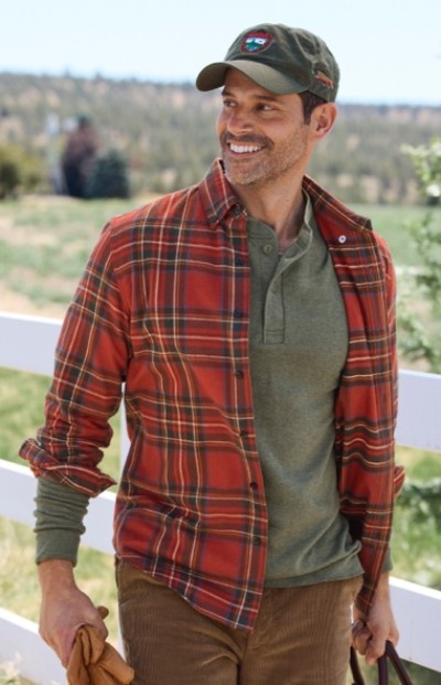 Man in red plaid shirt over green henley and brown pants holds gloves and bag, standing by white fence.