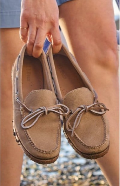 A hand holding tan leather moccasins with bow detailing, shown against an outdoor, sunlit background.