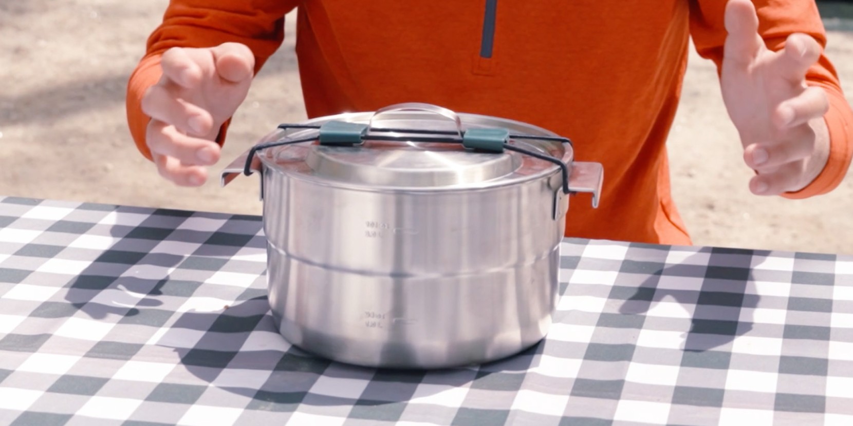 A person holds a metal pot with a lid on a checkered tablecloth.