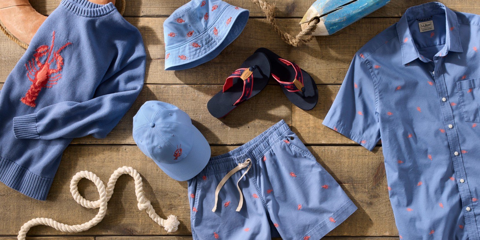 Flat lay of blue lobster-print sweater, shirt, shorts, hat and flip-flops arranged on a wooden surface. 