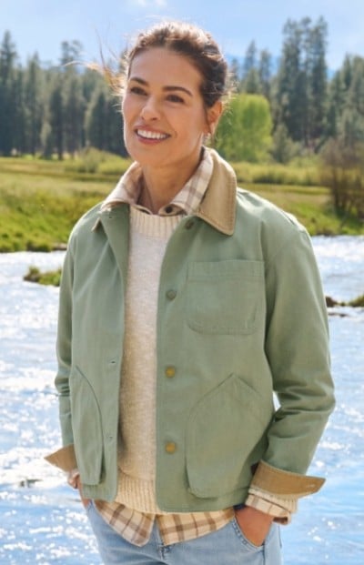 Woman in a green jacket, beige sweater, and plaid shirt stands by a river with trees in the background.