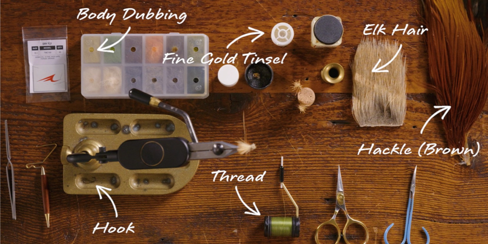 Fly tying tools and materials including body dubbing, fine gold tinsel, elk hair, hackle, hook, thread on a wooden surface.
