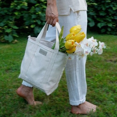 Waist-down shot of someone walking barefoot through the grass carrying a tote bag with fresh flowers.