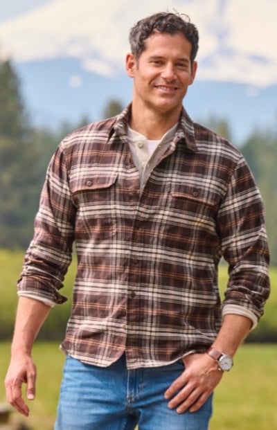 Person in brown plaid shirt and blue jeans stands outdoors, with a mountain and trees in the background.