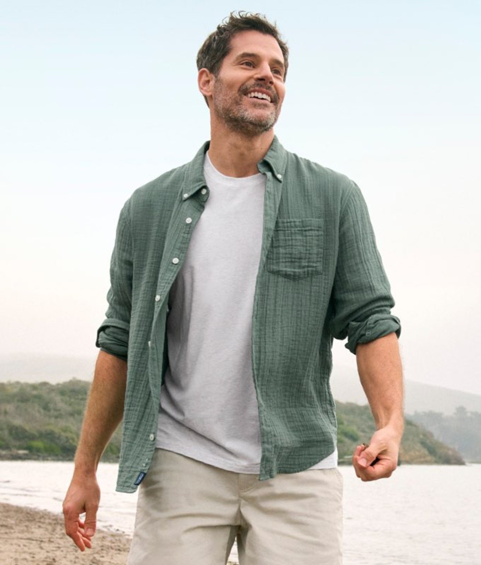 Person walking outside in a green button-up shirt over a white tee, with a waterfront city view behind.