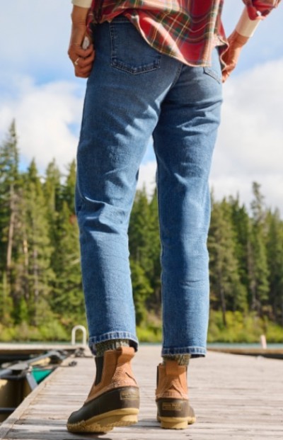 A woman wearing jeans and boots stands on a wooden dock with hands on hips, facing away. Trees and a cloudy sky fill the background.