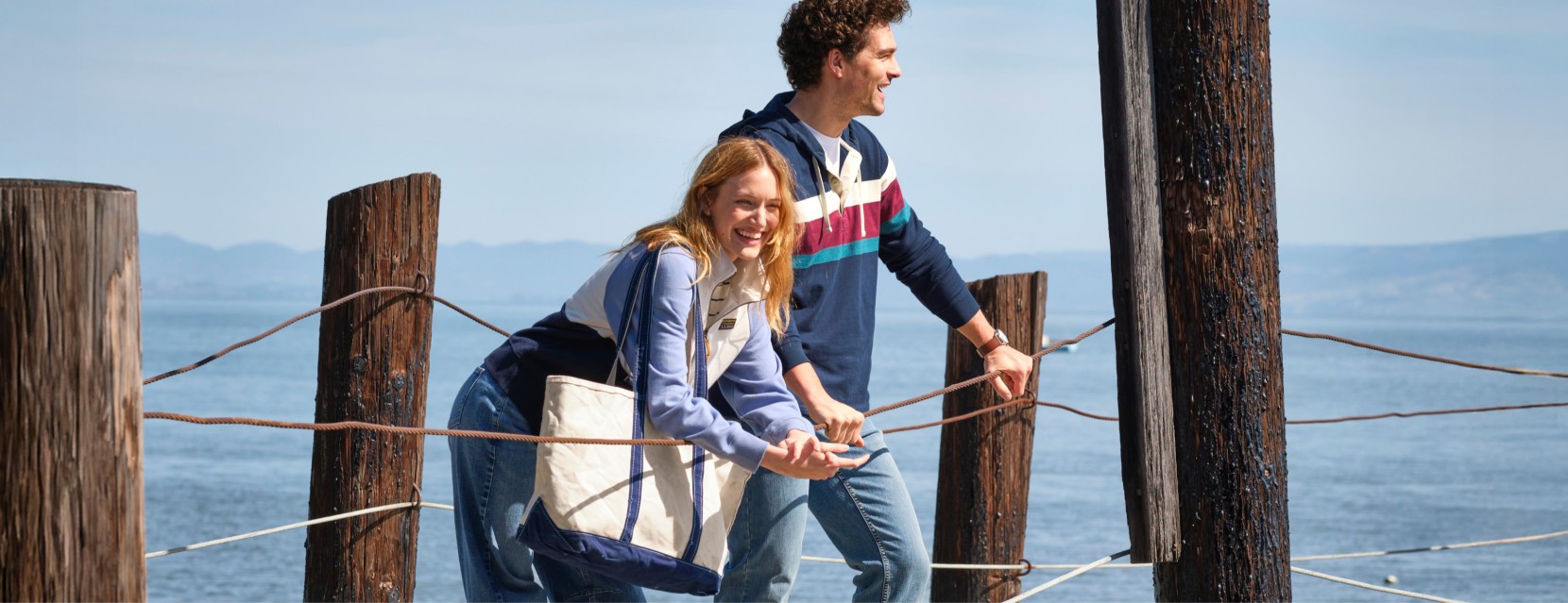 Two people stand on a waterfront pier, leaning on rope rails with calm water and distant hills behind them.