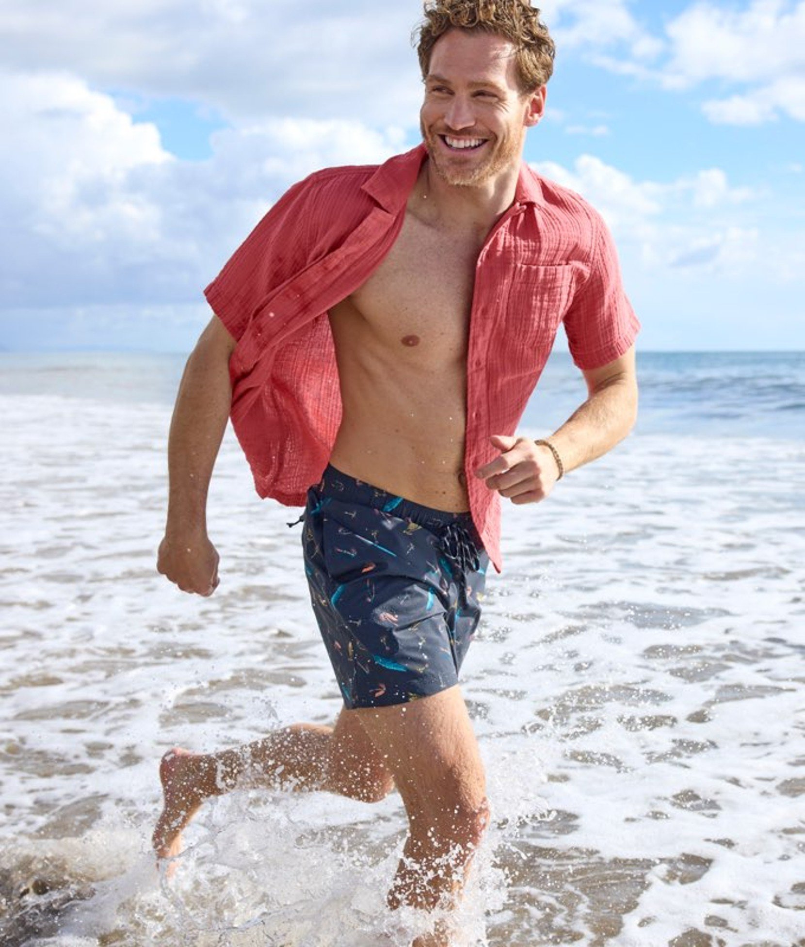 Person wearing swim shorts and a jacket, walking along the shoreline with waves nearby.