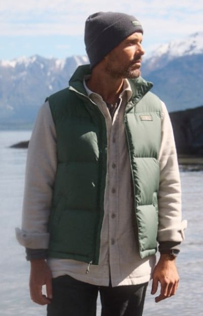Person wearing layers under a green insulated vest and knit hat stands beside a lake and mountains.