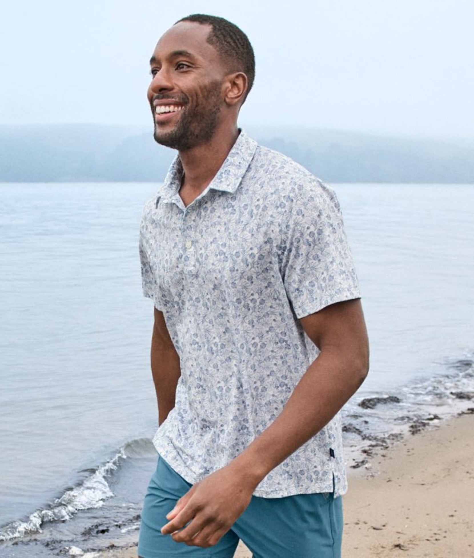 Person wearing a patterned short-sleeve shirt by the shoreline, with waves and blue sky visible.