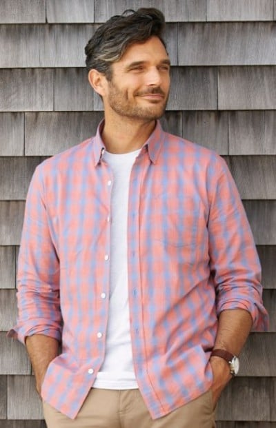 A man wearing a pink and blue checkered button-down shirt layered over a white tee, in front of a clapboard wall.