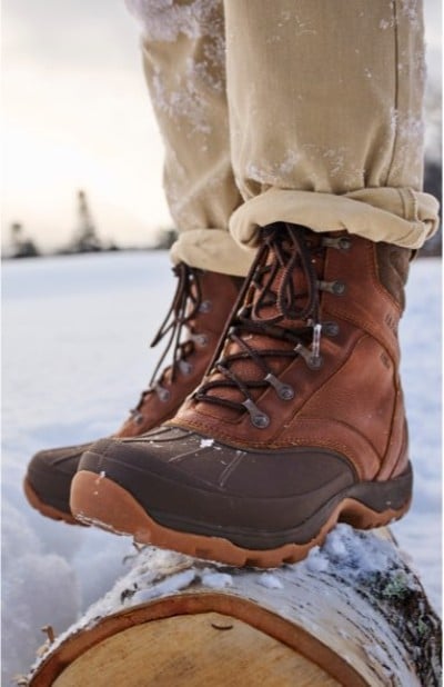Close up of a person balanced on a snowy log in a winter landscape wearing brown boots.