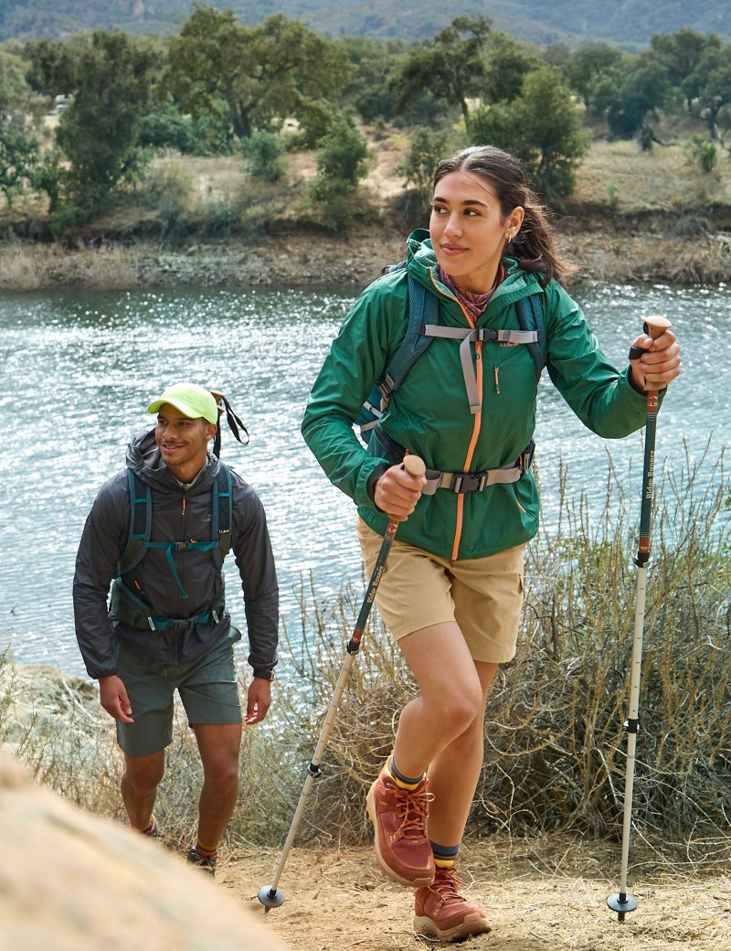 A man and woman wearing day packs hike up a trail, one carrying and one using hiking poles.