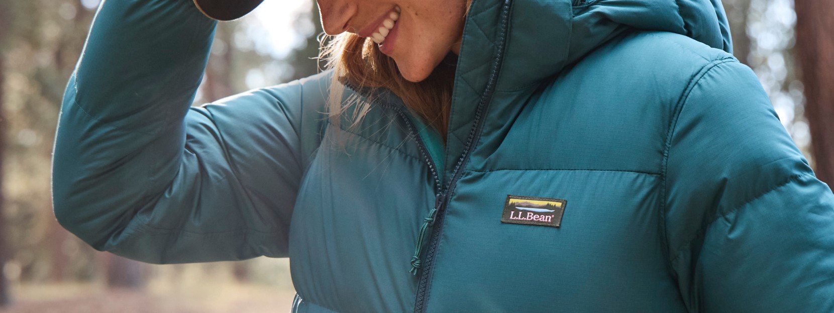 Woman in teal L L Bean puffer jacket smiling and holding hat brim, with a blurred forest background. 