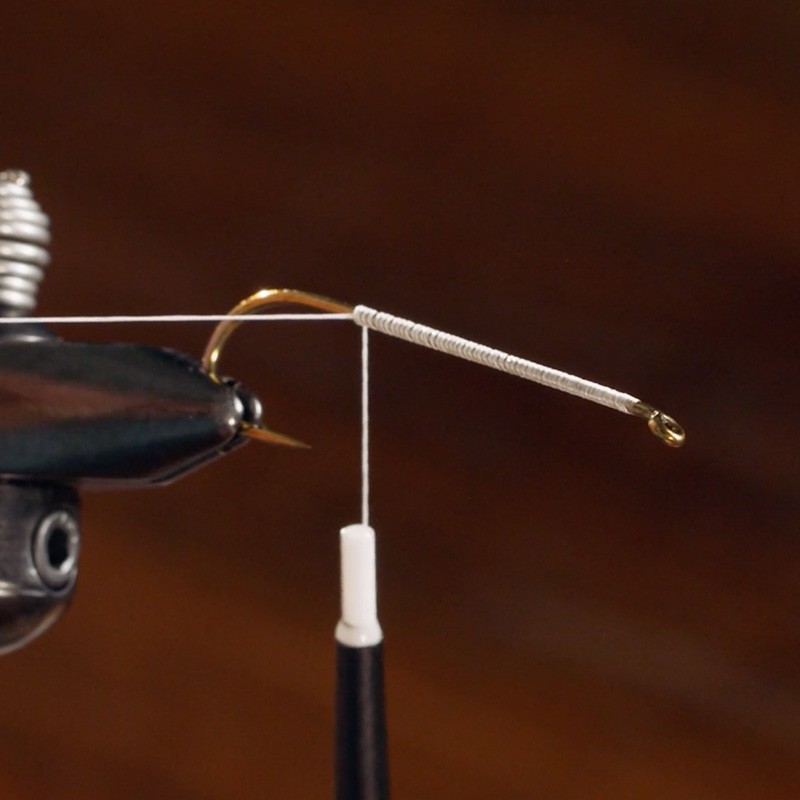 Close-up of a fly fishing hook being wrapped with white thread on a vise against a brown background.